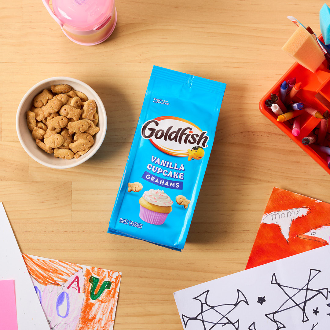 Goldfish Vanilla Cupcake Grahams with a bowl of graham crackers, children's artwork, and craft supplies on a wooden table.