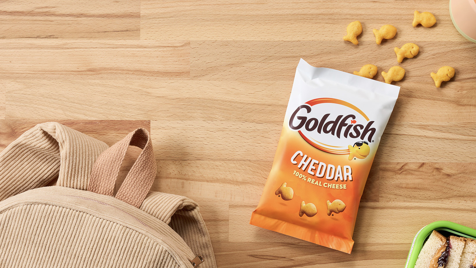 Goldfish® Cheddar crackers bag on a wooden table with a backpack, lunch container, and scattered crackers