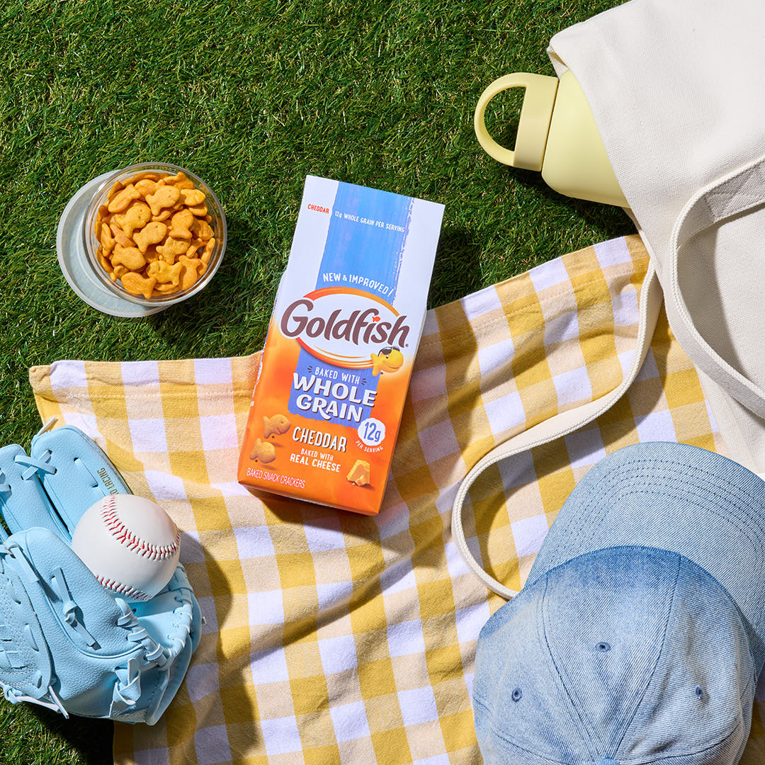 Goldfish Whole Grain Cheddar crackers with a baseball glove, baseball, and picnic blanket on a grassy field.
