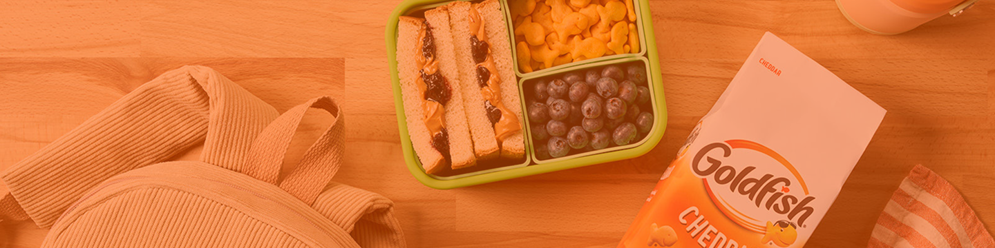 Packed lunch with a peanut butter and jelly sandwich, fruit, and Goldfish Cheddar crackers.