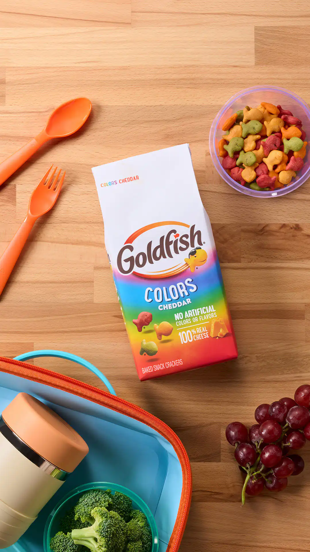 Goldfish Colors Cheddar crackers paired with grapes, broccoli, reusable lunch containers, and utensils for a packed lunch.